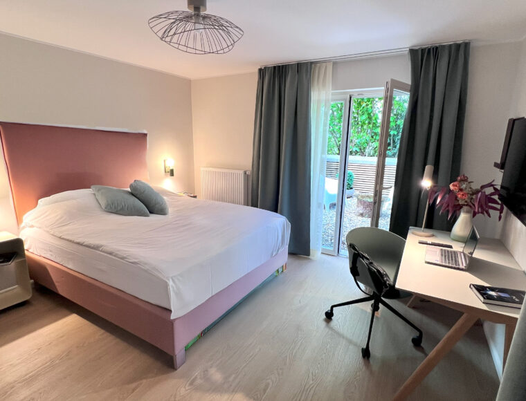 A bedroom with a double bed, pink headboard, and white linens by Keyinn Serviced Residences. Joined by a desk with a laptop, flowers & small garden.