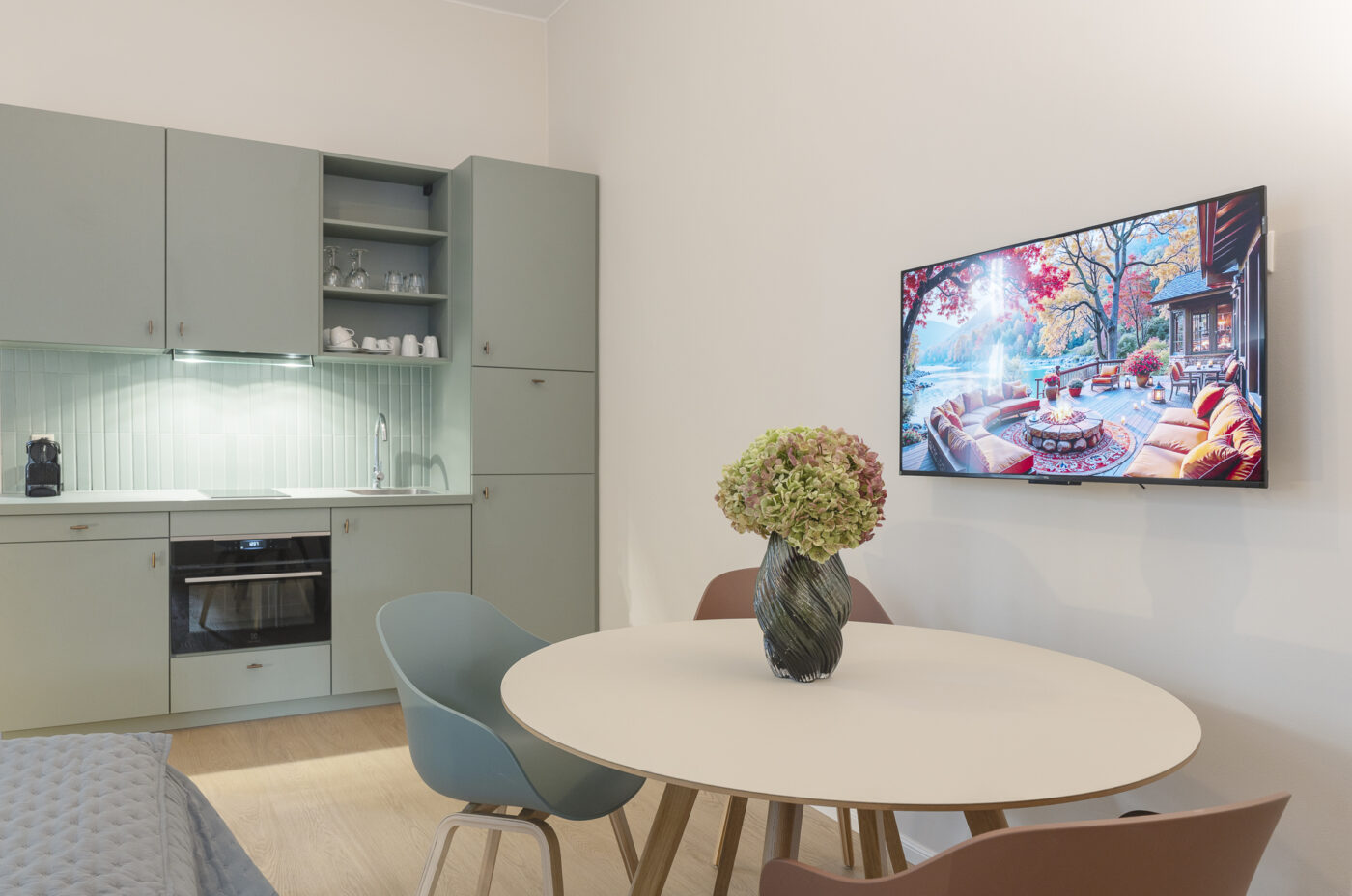 Keyinn Serviced Residences Belair kitchen with pale green cabinets and a round dining table with a floral centerpiece. The space features a wall-mounted TV and a bright, modern design.