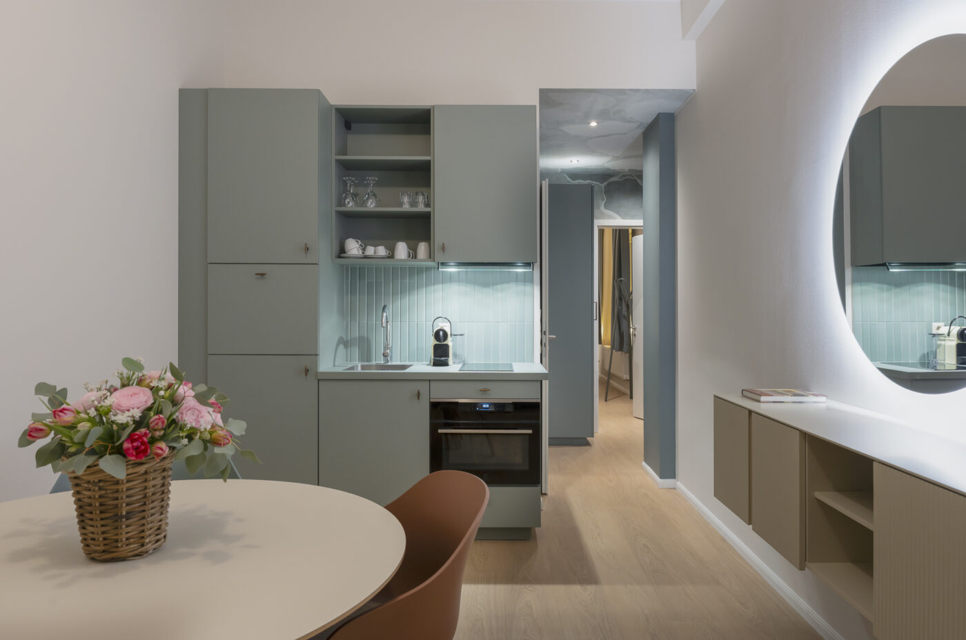 Keyinn Serviced Residences Belair kitchen with muted green cabinets and a round dining table with flowers. The space features a glowing circular mirror and a clean, modern aesthetic.