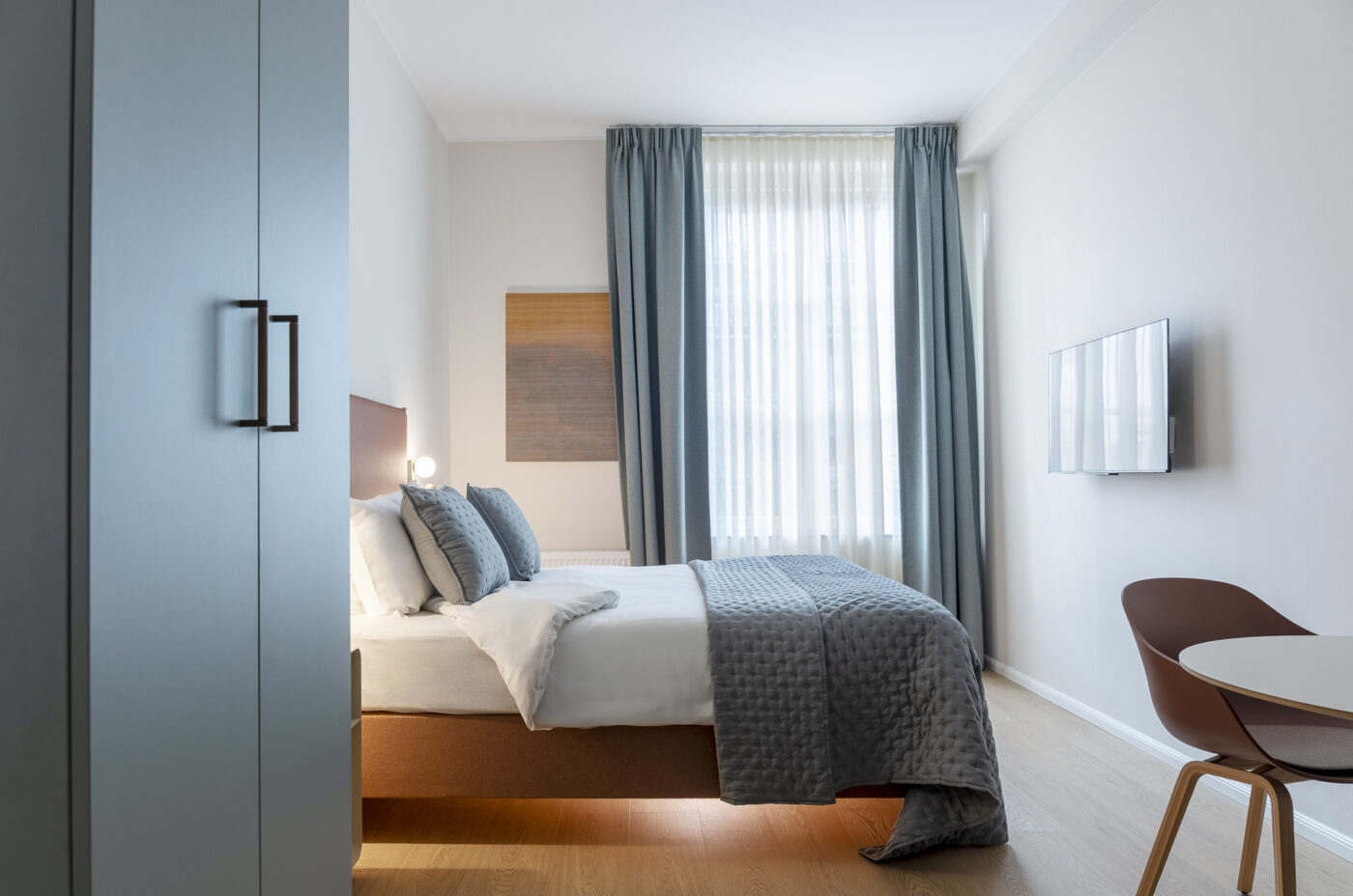 Keyinn Serviced Residences Belair bedroom with grey bedding, wall-mounted tv, and a window with white and blue-grey curtains.