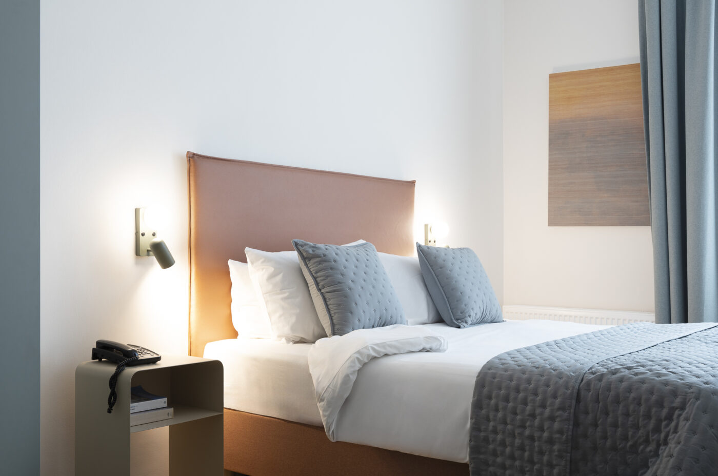 Keyinn Serviced Residences Belair room with blue-grey curtains, bedsheets & walls. Also features a small beige bedside table, with a telephone and books.