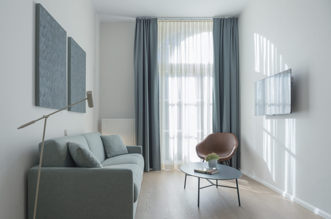 Keyinn Serviced Residences Belair living room with a teal sofa, circular coffee table, and a brown accent chair. The space features abstract wall art, a mounted TV, and large windows with matching teal curtains.