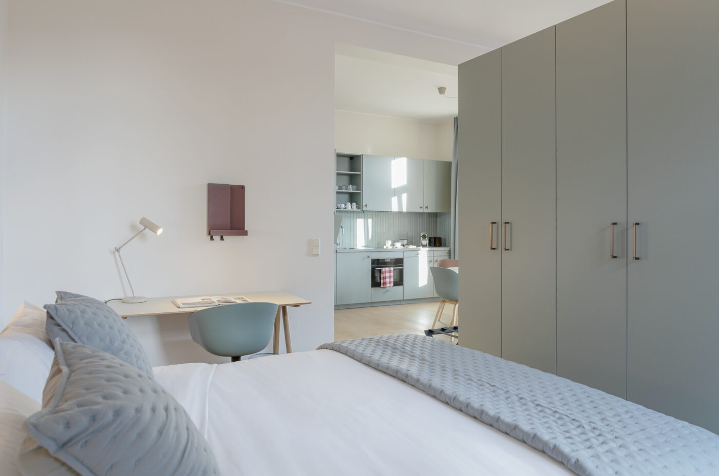 Keyinn Serviced Residences Belair bedroom with teal pillows, a desk, and a modern lamp. The space features an open view into a minimalist kitchen with a soft colour palette throughout.