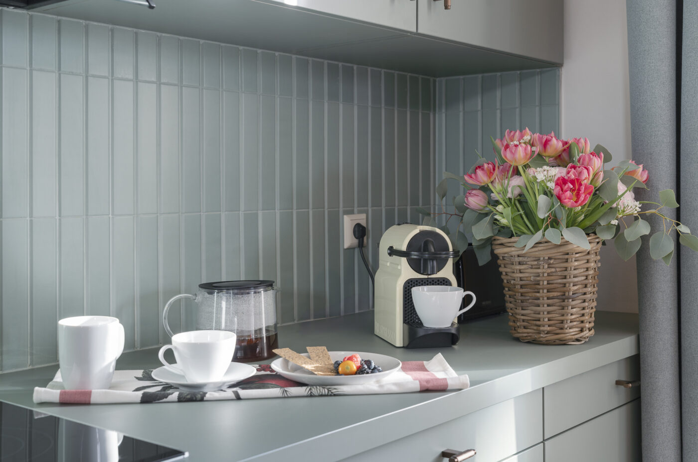 Keyinn Serviced Residences Belair kitchen counter with pastel green tiles, a coffee machine, and a glass jug. The space features a plate of berries, biscuits, and a basket of pink flowers.