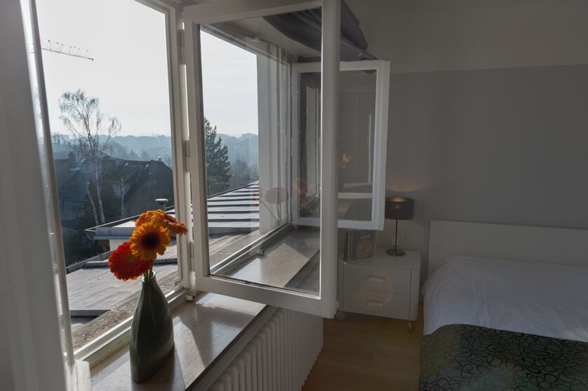 A Keyinn Serviced Residences studio apartment featuring an open window that showcases a scenic, misty view of distant trees and rooftops. A vase of vibrant flowers sits on the windowsill.