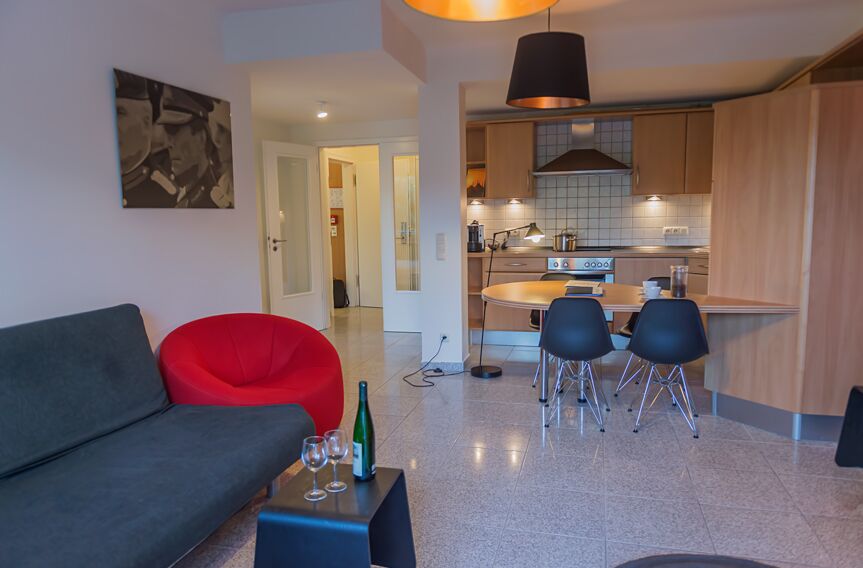 Keyinn Serviced Residences Parc de Merl living and dining space with a black sofa, red chair, and glassware.