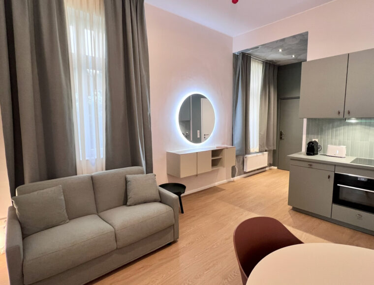 Keyinn Serviced Residences Belair living room with a grey sofa and an illuminated round mirror. The space features a compact kitchen with gray cabinets and a modern, minimalist design.