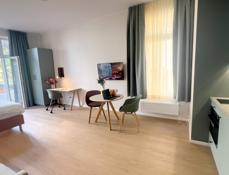 A Keyinn Serviced Residences studio apartment with light wood flooring, featuring a bed, a small dining table with two chairs, a wall-mounted TV, and a kitchenette.