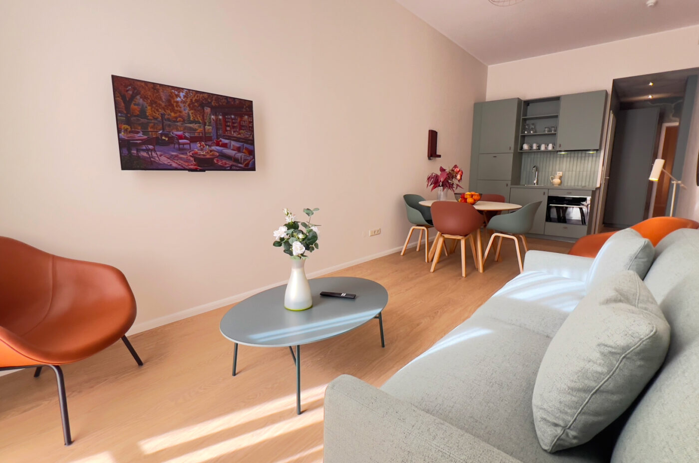 Keyinn Serviced Residences apartment living room featuring a light gray sofa and a bold orange armchair on wooden flooring. A floral centrepiece rests on a gray coffee table, with a glimpse of the kitchen in the background.