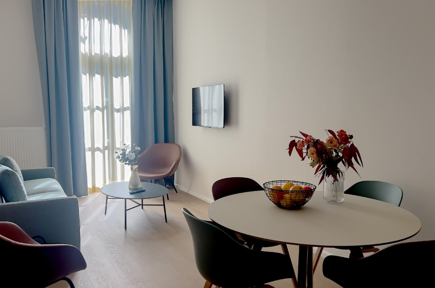 Keyinn Serviced Residences Belair living room featuring a blue sofa, round coffee table, and a wall-mounted TV. The space includes a dining table with a floral centerpiece and fruit bowl, set against a clean, modern design.