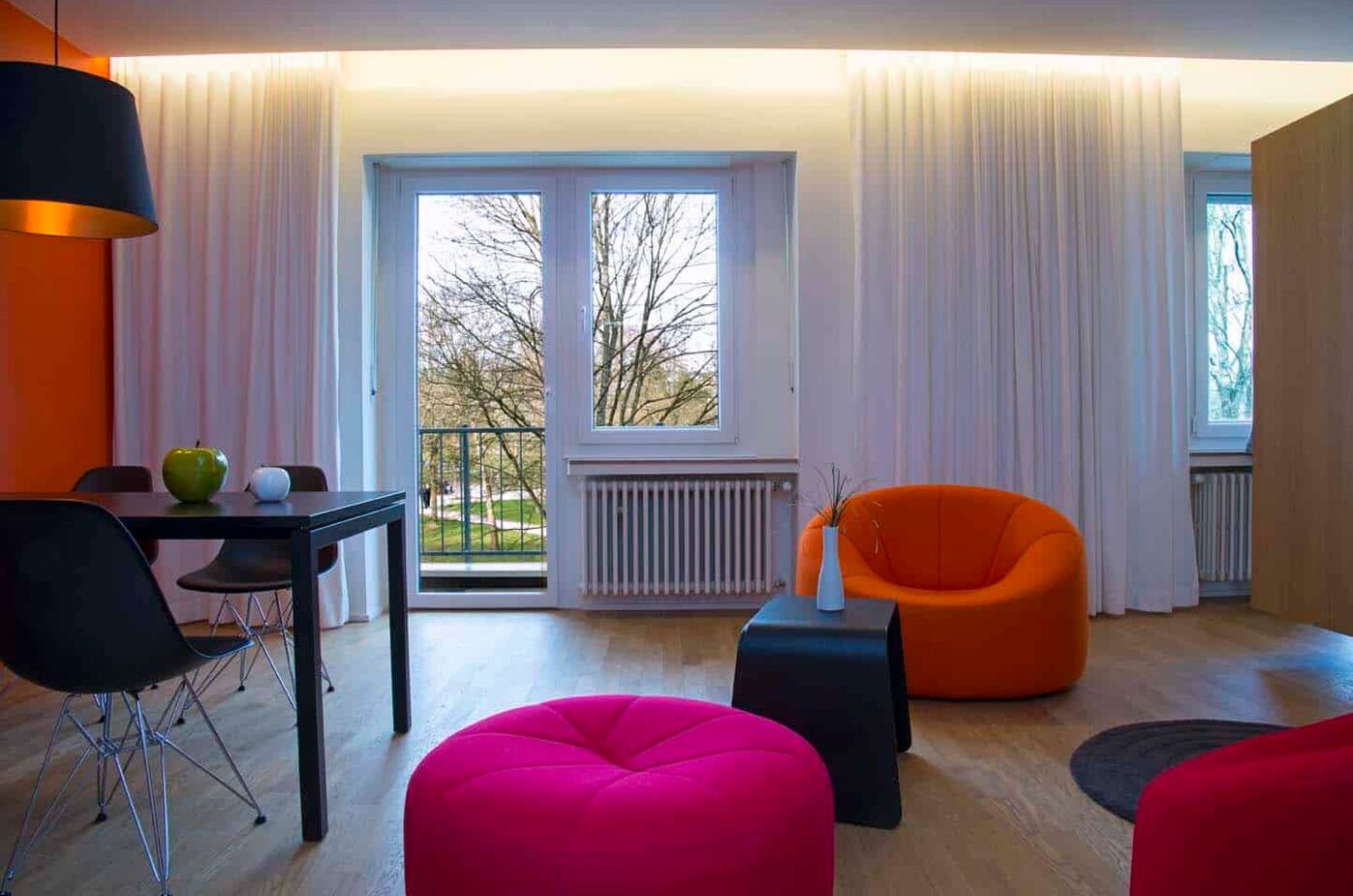 Keyinn Serviced Residences Parc de Merl living room with orange, pink & red seating, and a black dining table joined by a balcony.