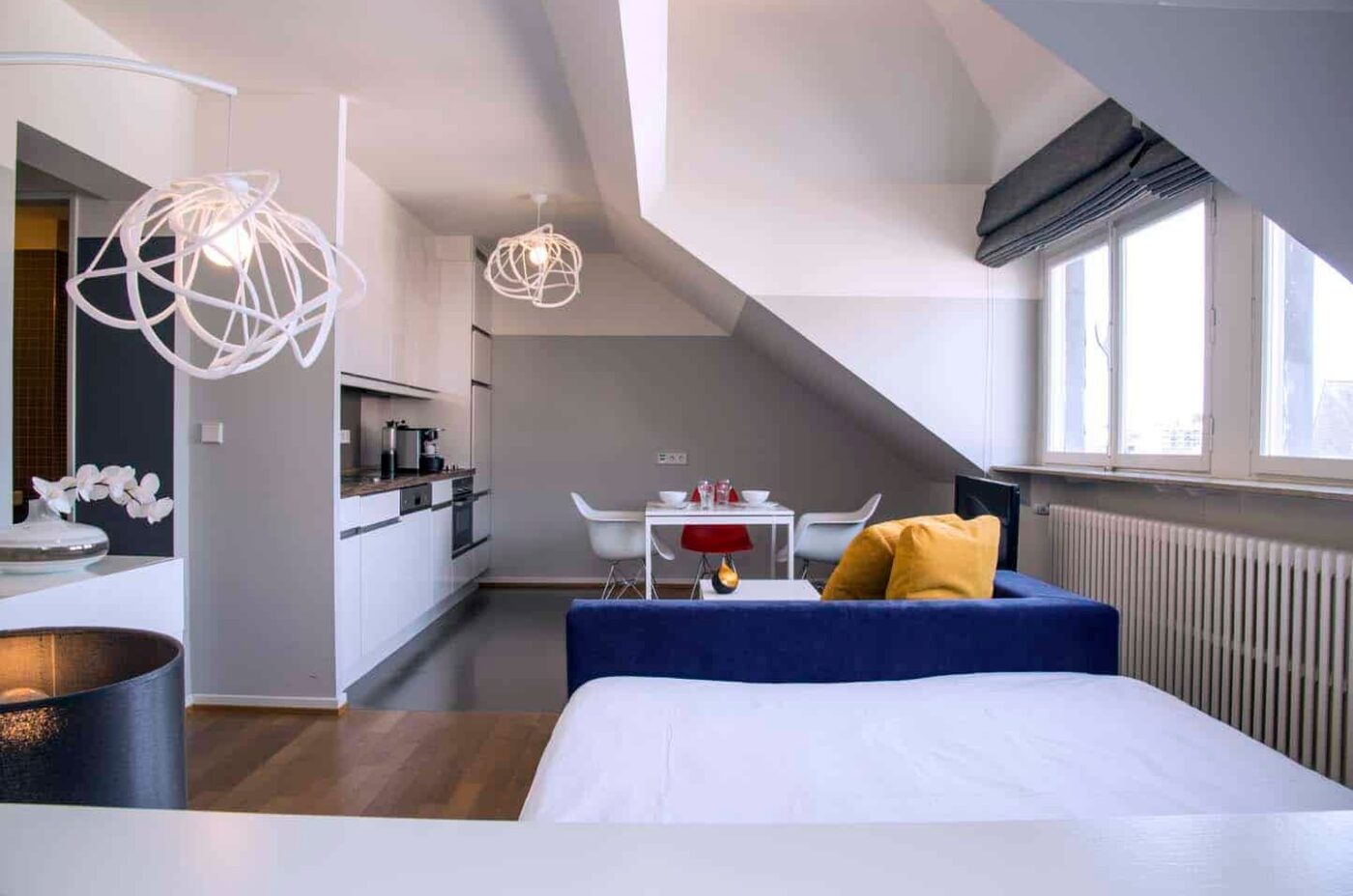 Keyinn Serviced Residences studio apartment with a sloped ceiling, featuring a blue sofa, white kitchen, and dining area. With pendant lights hanging from the ceiling.