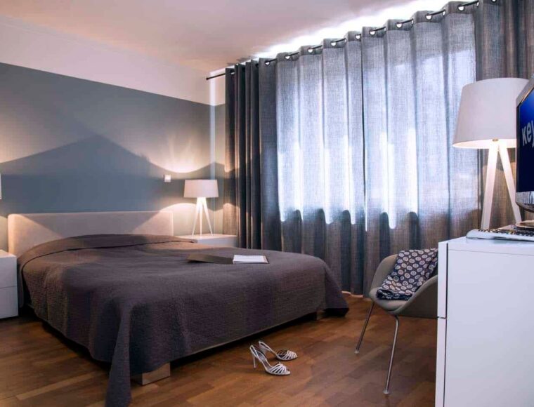 A bedroom at Keyinn Serviced Residences featuring a gray bedsheets and curtains, with blue themed wall. White lamps and a TV positioned on a dresser.