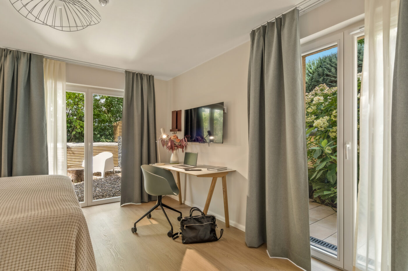 Modern room at Keyinn Serviced Residences with a beige bed, light wooden floor, and a desk with a laptop. Large windows with gray curtains open to a lush garden, creating a serene atmosphere.