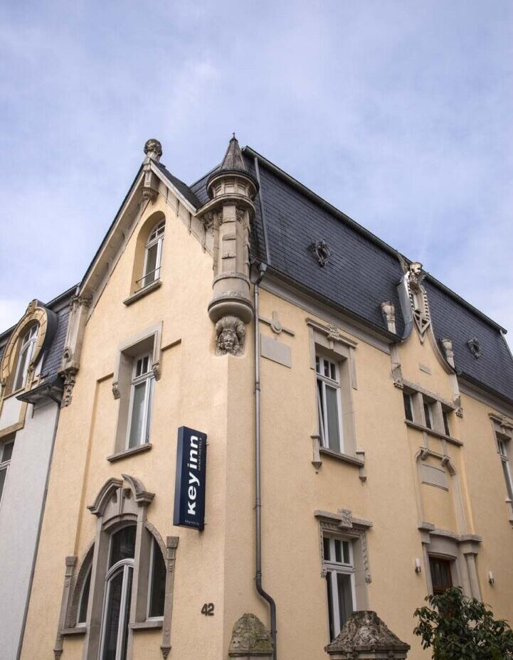 Historic beige building with ornate details, arched windows, and a turret under a cloudy sky. A sign reads 