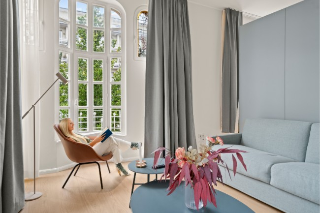 A serene, bright living room at Keyinn Serviced Residences, with tall arched windows and gray curtains. A person is sitting on a brown chair reading.