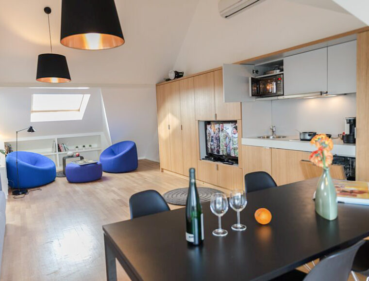 Modern open-plan apartment at Keyinn Serviced Residences with wooden floors, integrated kitchen, dining table set for two, TV, and two blue chairs near a window.