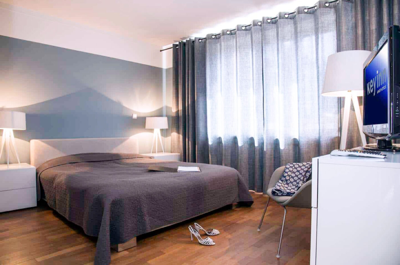 A sophisticated bedroom at Keyinn Serviced Residences styled in grey tones. The space features a double bed, soft illumination from two bedside lamps, and grey curtains.