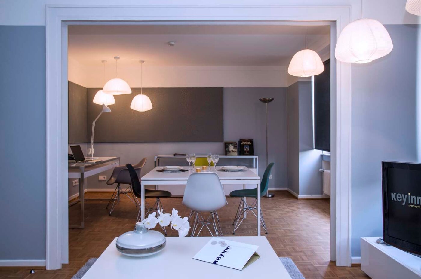 A modern dining & living area at Keyinn Serviced Residences features a dining table set for four, contemporary chairs, stylish pendant lights, & a TV.