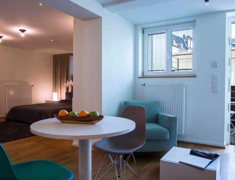 A Keyinn Serviced Residences apartment interior with natural light. Features a round table with a fruit bowl, seating area, bed in the background & patio door open.