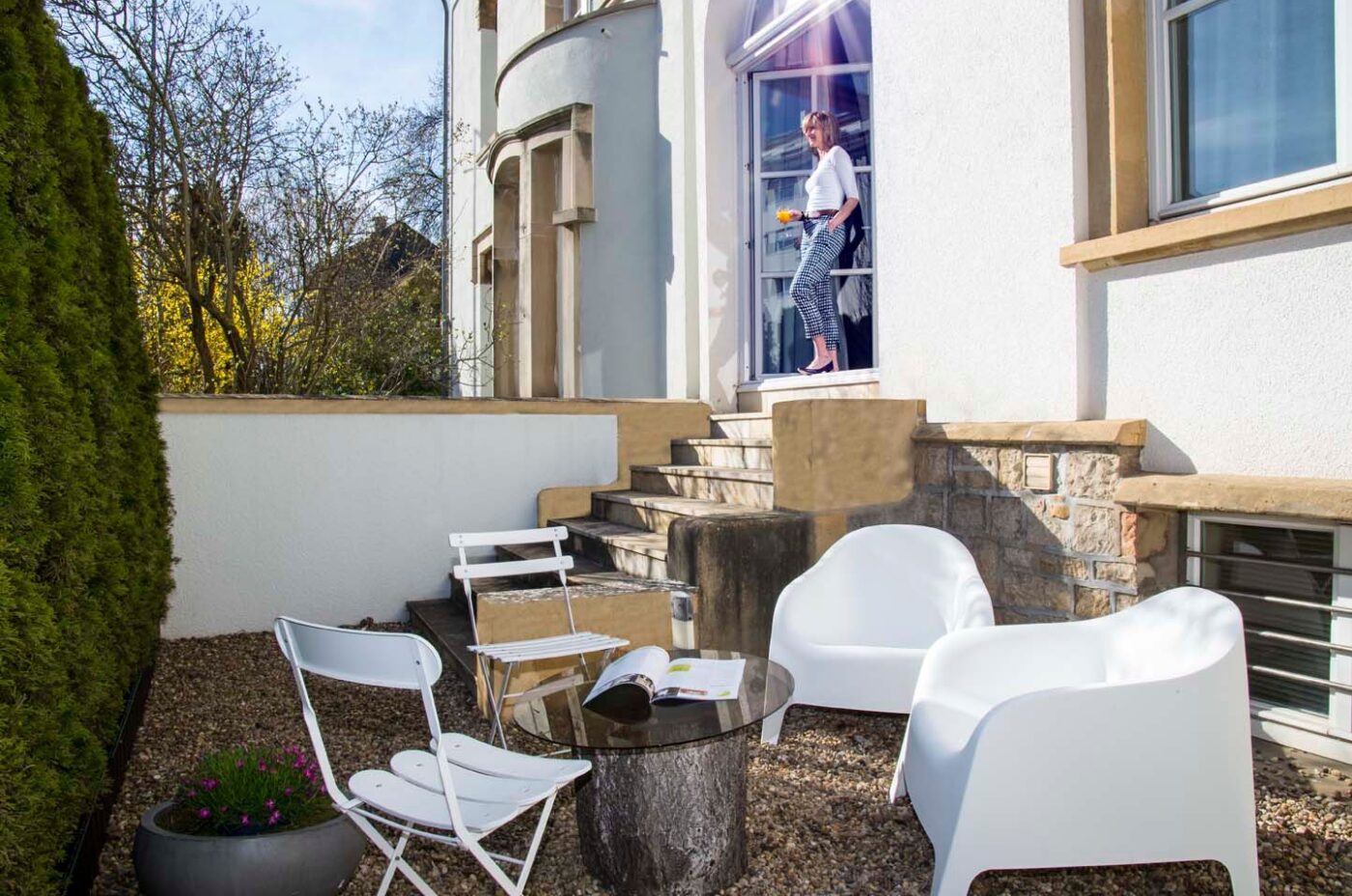 Sunlit patio with modern white chairs and a glass-top table on gravel at Keyinn Serviced Residences. A person stands on stairs holding a tray near an arched doorway, conveying a relaxed, inviting atmosphere.