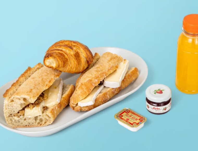 Breakfast arrangement on a vibrant light blue surface with baguette brie sandwiches, a croissant, orange juice, and jam.