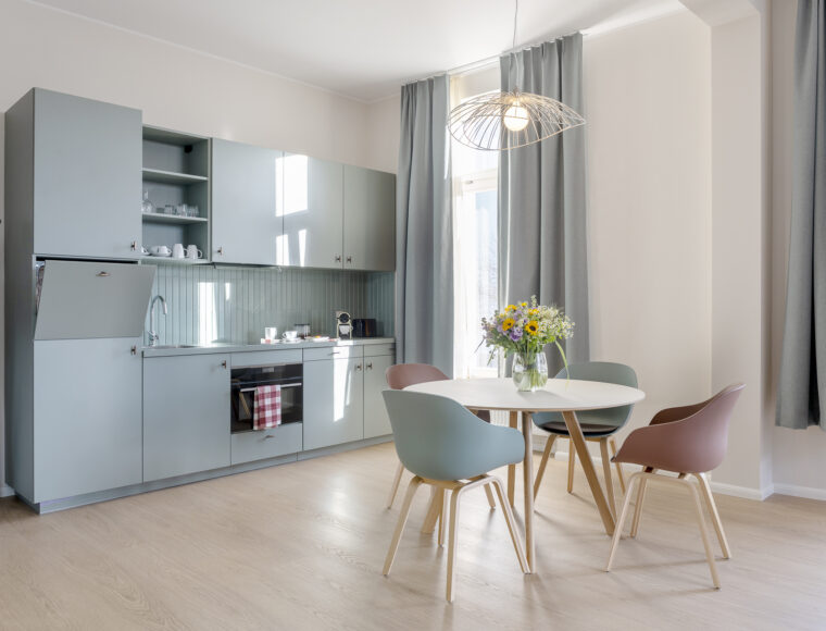KeyInn Serviced Residences Cuisine moderne avec des armoires bleu pâle, un four encastré et des étagères ouvertes. Une table à manger ronde avec quatre chaises aux couleurs pastel et un vase de fleurs se trouve à proximité. De grandes fenêtres avec des rideaux gris laissent entrer la lumière naturelle.