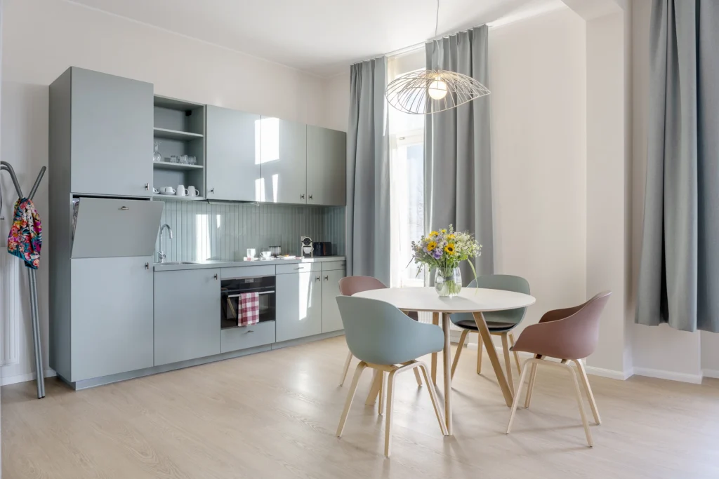 KeyInn Serviced Residences Modern kitchen with light blue cabinets, built-in oven, and a dining area featuring a round table with four colorful chairs. A bouquet of flowers sits on the table; large windows let in natural light.